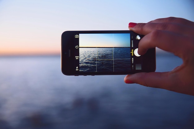 Przycięta ręka kobiety trzymająca smartfon podczas fotografowania morza w zmierzchu