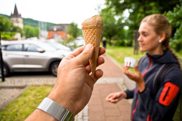 Zdjęcie przycięta ręka mężczyzny trzymającego stożek z lodem przeciwko kobiecie stojącej na chodniku w tle