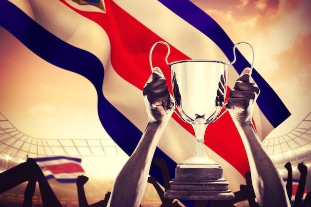 Przycięta ręka sportowca trzymającego trofeum na dużym stadionie piłkarskim pod pochmurnym błękitnym niebem