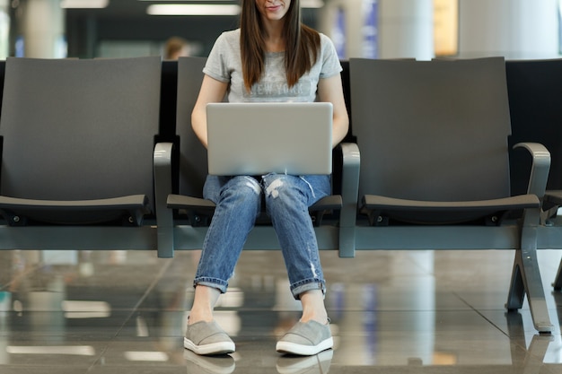 Przycięte młoda uśmiechnięta podróżnik turystyczna kobieta pracuje na laptopie podczas oczekiwania w holu na międzynarodowym lotnisku