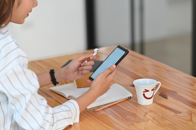 Przycięte ujęcie kobiety w pasiastej koszuli, trzymając w ręku biały pusty ekran smartfona i bezprzewodowe słuchawki w ręku, siedząc przy drewnianym biurku.