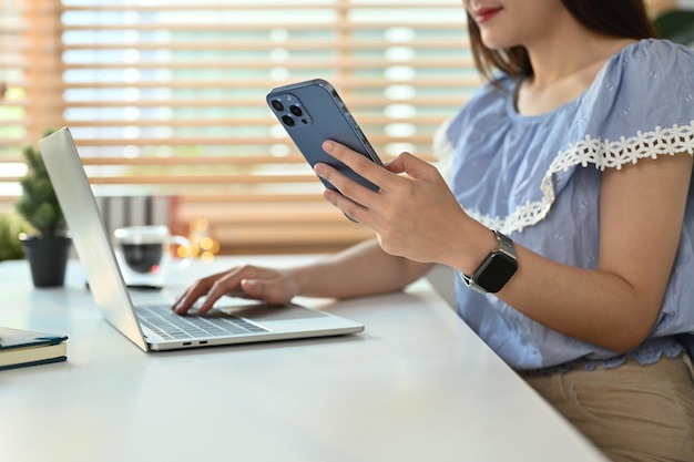 Przycięte ujęcie przedsiębiorczej kobiety rozmawiającej z klientem online na smartfonie i korzystającej z laptopa w swoim miejscu pracy