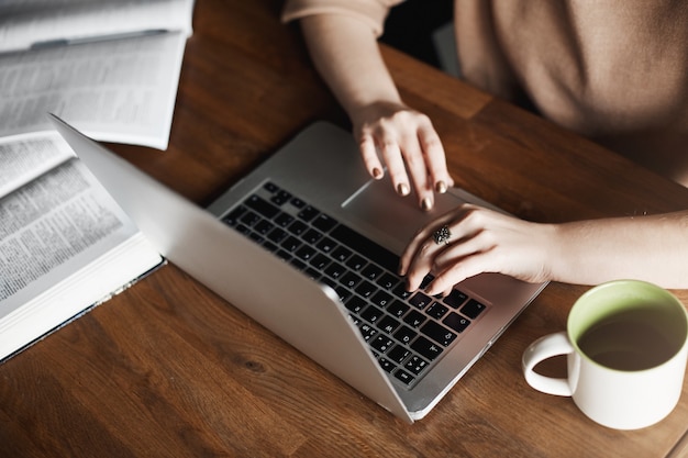 Przycięte ujęcie skoncentrowanej kobiety siedzącej w biurze przy stole, pisząc na klawiaturze laptopa, pijąc herbatę