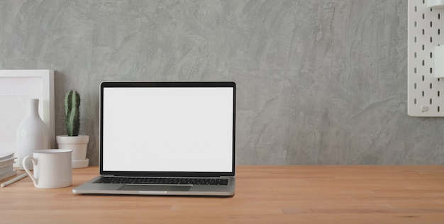 Przycięte ujęcie stylowego miejsca pracy z otwartym laptopem z pustym ekranem i materiałami biurowymi na drewnianym stole i poddaszu szarej ścianie