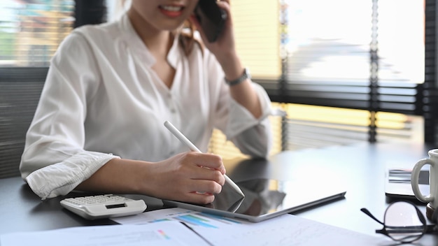 Przycięte zdjęcie kobiecego doradcy finansowego za pomocą cyfrowego tabletu i rozmawiającego przez telefon komórkowy z klientem