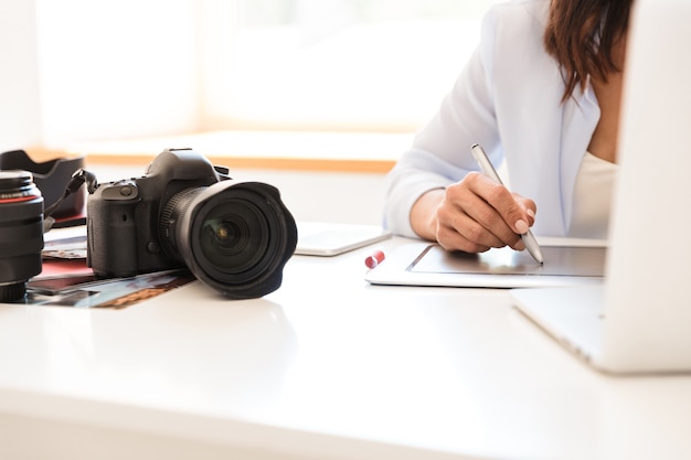 Przycięte zdjęcie kobiety fotograf w biurze siedzącej przy stole z aparatem pracującym przez tablet graficzny retuszujący zdjęcia.