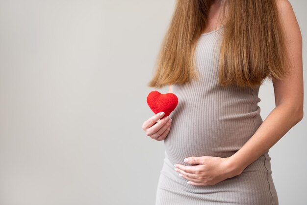 Przycięte zdjęcie kobiety w ciąży noszącej obcisłą sukienkę trzymającą miękkie czerwone serce na szarym tle Pregnan