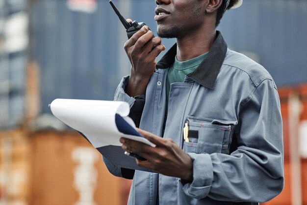 Przycięte zdjęcie młodego czarnego mężczyzny używającego krótkofalówki podczas pracy w dokach wysyłkowych z kontenerami