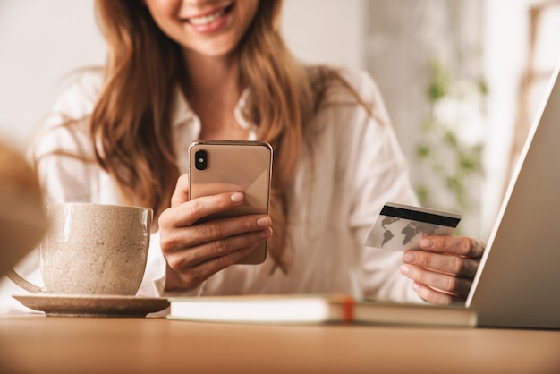 Przycięte zdjęcie młodej wesołej kobiety imbir siedzieć w pomieszczeniu w biurze przy użyciu komputera przenośnego i telefonu komórkowego, trzymając kartę kredytową.