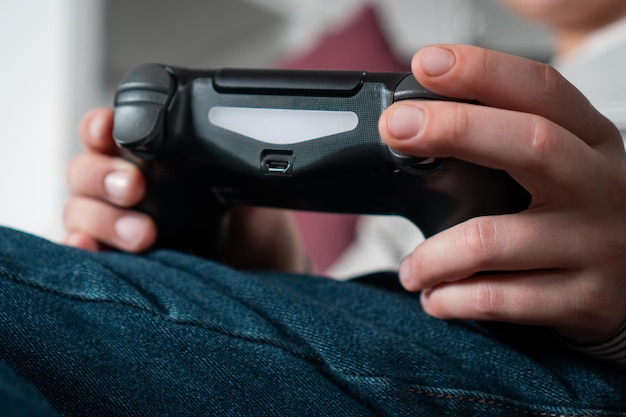 Przycięte zdjęcie nastoletniego chłopca siedzącego w domu trzymającego czarny joystick kontrolera gier grających w gry wideo Widok z tyłu