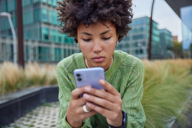 Przycięte Zdjęcie Poważnej Młodej Kobiety Z Kręconymi Włosami Przegląda Telefon Komórkowy Wysyła Wiadomości Tekstowe Nosi Zielony Sweter Pozuje Na Rozmytym Tle Miejskim Nowoczesne Technologie I Koncepcja Stylu życia