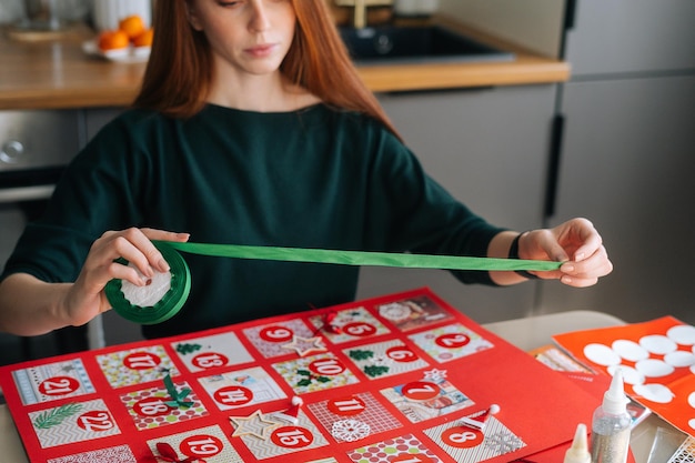 Przycięte zdjęcie skoncentrowanej rudej młodej kobiety używającej świątecznej wstążki do oprawienia tablicy z kopertami z kalendarza adwentowego w domu