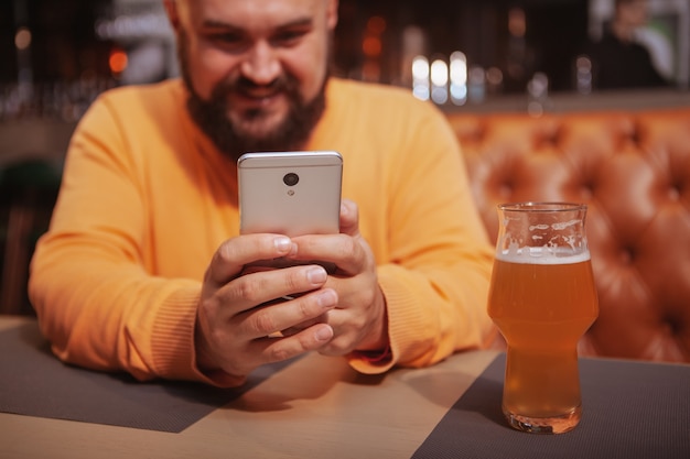 Przycięte zdjęcie szczęśliwego brodatego mężczyzny, uśmiechając się, za pomocą swojego inteligentnego telefonu w pubie piwa