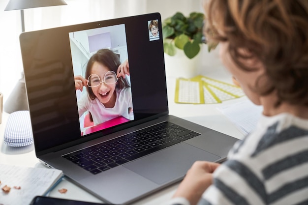 Przycięty dzieciak wykonujący rozmowy wideo na nowoczesnym laptopie z pozytywną dziewczyną w okularach przy stole z notatnikiem w jasnym pokoju
