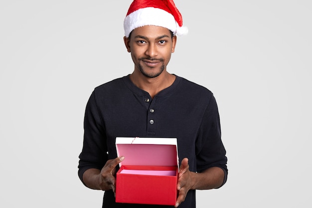 Przycięty izolowany, ciemnoskóry młody mężczyzna nosi czapkę Mikołaja, ma otwarte czerwone pudełko, sugeruje kupienie prezentu na Boże Narodzenie, stoi na białym. Koncepcja wakacje