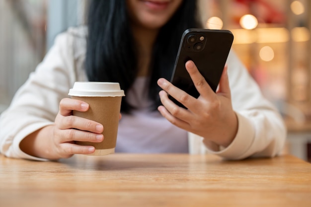 Przycięty obraz azjatyckiej kobiety z filiżanką kawy i smartfonem siedzącej w kawiarni