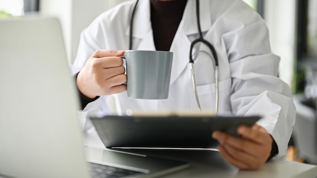 Przycięty obraz inteligentnej lekarki popijającej kawę i czytającej przypadki medyczne w schowku