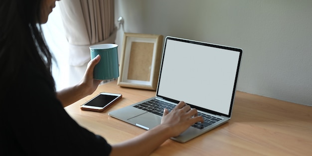 Przycięty obraz kobiety używa białego laptopa z pustym ekranem, siedząc w salonie.