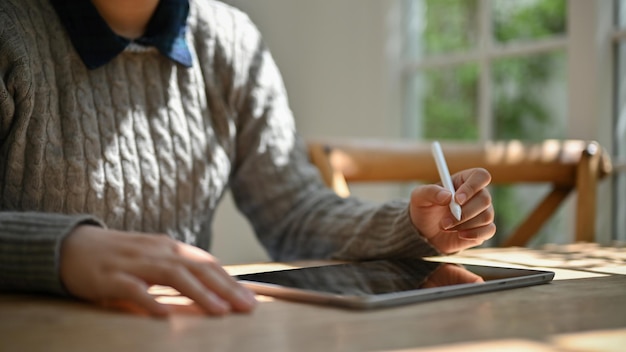 Przycięty obraz kobiety w przytulnym swetrze pracującej nad swoimi zadaniami na tablecie cyfrowym
