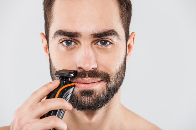 Przycięty obraz przystojny brodaty mężczyzna do golenia z elektryczną maszynką do golenia na białym tle nad białym