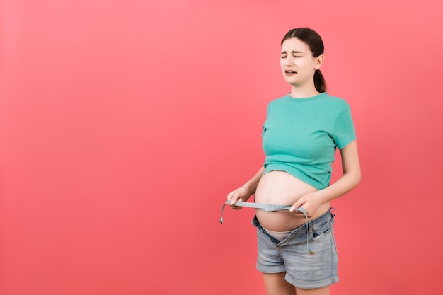 Przycięty obraz spodziewającej się matki w rozpiętych dżinsach, mierzącej swój ciężarny brzuch za pomocą taśmy mierniczej na kolorowym tle Pomiar centymetra