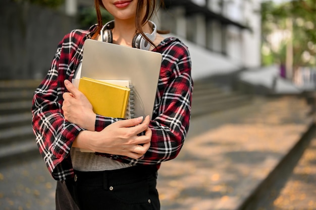 Zdjęcie przycięty obraz studentki college'u trzymającej laptopa i książkę szkolną stojącą na zewnątrz