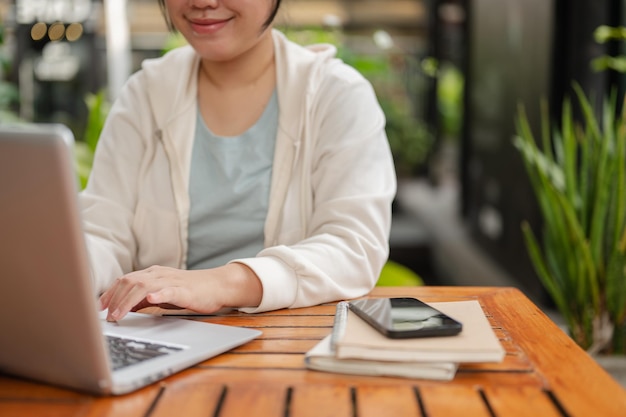 Przycięty obraz szczęśliwej azjatyckiej kobiety w casualowym ubraniu pracuje na swoim laptopie w przestrzeni zewnętrznej