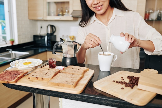 Przycięty Obraz Uśmiechniętej Młodej Azjatki, Która Rano Nalewa śmietankę Do Kawy Podczas Robienia Sobie śniadania