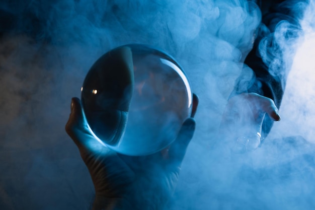 Przycięty widok męskiej dłoni z kryształową kulą i kobiecej dłoni z dymem wokół ciemnoniebieskiego