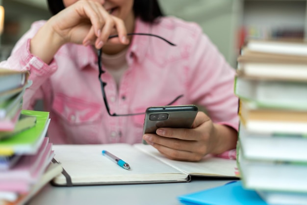 Przycięty Widok Uśmiechniętej ładnej Kaukaskiej Kobiety Trzymającej Okulary Za Pomocą Telefonu Komórkowego I śmiejącej Się Podczas Odrabiania Lekcji W Bibliotece