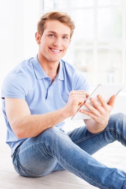 Przyglądam się jego zupełnie nowemu tabletowi. Przystojny młody mężczyzna pracuje na cyfrowym tablecie i uśmiecha się siedząc na podłodze w swoim mieszkaniu