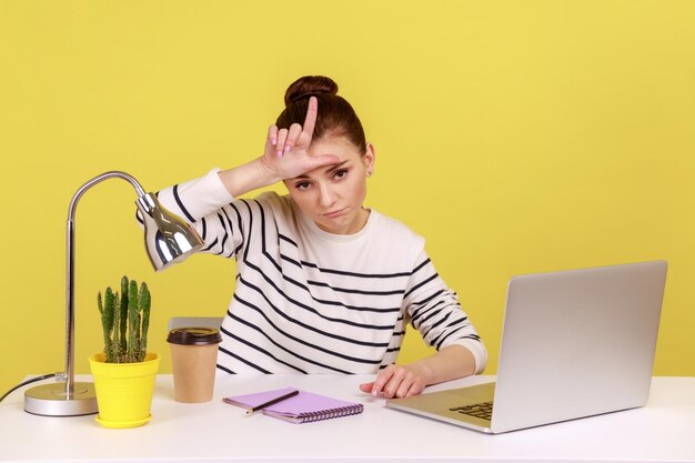 Przygnębiona kobieta siedzi w miejscu pracy i pokazuje gest przegrany na ekranie laptopa