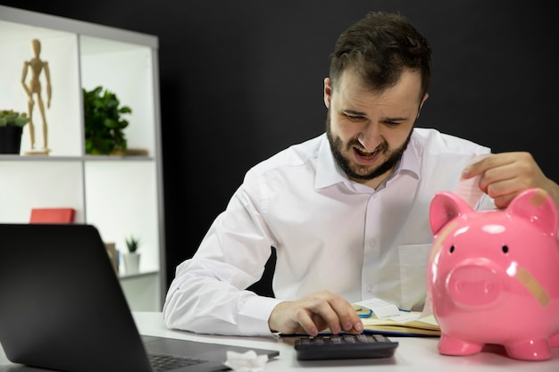 Przygnębiony biznesmen z łamaną prosiątko bankiem na biurku oblicza rachunki