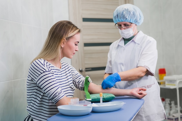 Przygotowanie do badania krwi z śliczną młodą blond kobietą przez kobietę lekarza w białym fartuchu mundur medyczny na stole w białym jasnym pokoju. Pielęgniarka przebija żyłę ramienia pacjenta pustą rurką z igłą.