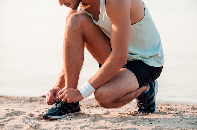 Przygotowanie do jego treningu. Przycięty obraz młodego mężczyzny wiążącego sznurowadła na butach sportowych podczas siedzenia na plaży