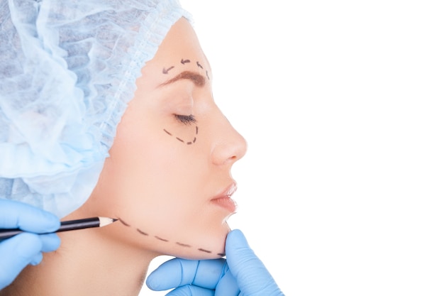 Zdjęcie przygotowanie pacjenta do zabiegu. widok z boku pięknej młodej kobiety w medycznym nakryciu głowy, trzymając zamknięte oczy, podczas gdy lekarz szkicuje jej twarz na białym tle