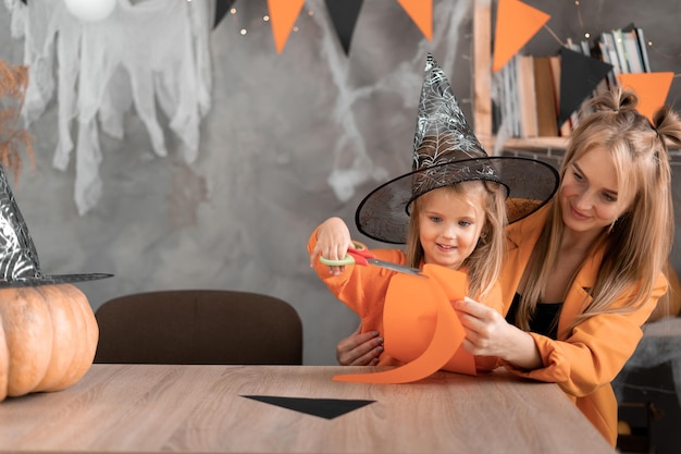 Zdjęcie przygotowując się do wakacji halloweenowa kobieta i córka siedzą w domu przy stole w kapeluszu wiedźmy i wycinają papierowe rękodzieła mama i dziecko dekorują pokój na święta