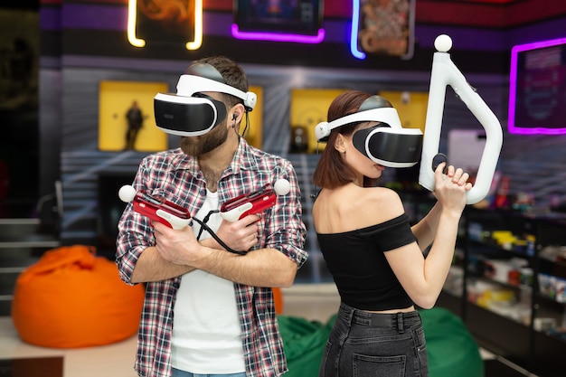 Przyjaciele, mężczyzna i kobieta używają na placu zabaw słuchawek wirtualnej rzeczywistości z okularami i kontrolerami ruchu dłoni oraz bronią.