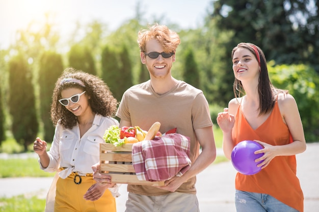Przyjaciele, rozrywka. Trzech młodych szczęśliwych przyjaciół w zwykłych ubraniach z piłką i koszem jedzenia spacerujących na pikniku w słonecznym zielonym parku
