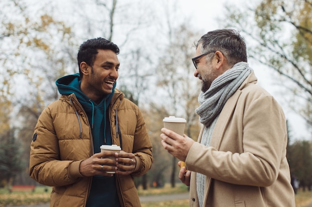 Przyjaciele seniora i młodego mężczyznę spacerują, rozmawiają i piją razem kawę w jesiennym parkuxA
