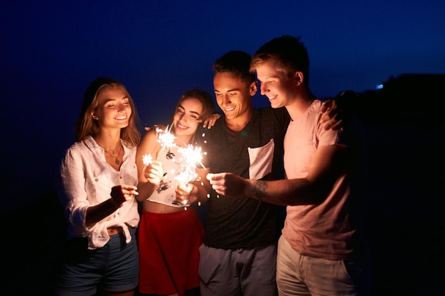 Przyjaciele Spacerują, Tańczą I Bawią Się Podczas Nocnej Imprezy Nad Morzem Przy światłach Bengalskich Brylantów