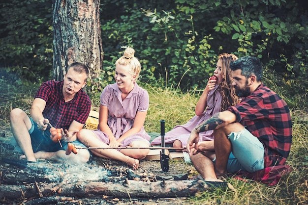 Przyjaciele urządzają piknik przy ognisku w lesie Mężczyźni i kobiety pieczą kiełbaski na ogniu Chłopaki i dziewczyny jedzą na kempingu Ludzie odpoczywają na letniej przyrodzie Kemping i gotowanie na wakacjach