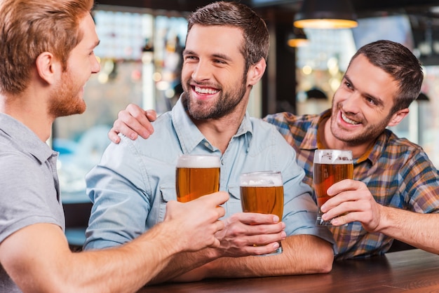 Przyjaciele w barze. Trzech szczęśliwych młodych mężczyzn w strojach codziennych rozmawiających i pijących piwo, siedząc razem przy barze?