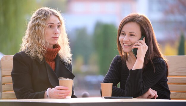 Przyjacielki Spędzają Czas Na Ulicznej Kawiarni Na świeżym Powietrzu, Podczas Gdy Jedna Dziewczyna Szczęśliwie Rozmawia Przez Telefon Komórkowy, A Druga Czeka Sfrustrowana Obraźliwym Zachowaniem Swojego Partnera.