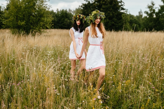 Przyjaźń kobiet. Nastrojowy portret dwóch pięknych słowiańskich dziewcząt w etnicznych sukienkach i wieńcu z kwiatami chodzącymi po naturze.