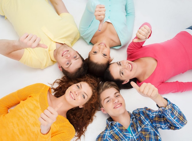 przyjaźń, młodość, gest i ludzie - grupa uśmiechniętych nastolatków leżących w kręgu na podłodze i pokazujących kciuki w górę