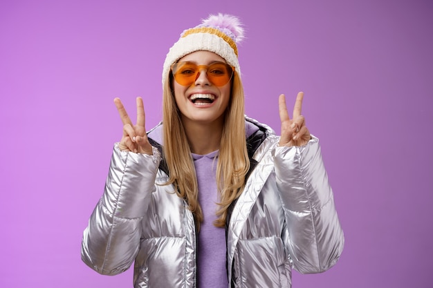 Przyjazna szczęśliwa optymistyczna piękna blond kobieta w okularach przeciwsłonecznych srebrna stylowa kurtka zimowa czapka pokazuje gest zwycięstwa pokoju, ciesząc się śnieżnymi wakacjami, wyjdź na zewnątrz hotelu ciesz się jazdą na nartach, fioletowe tło.