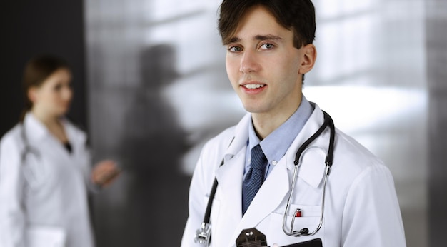 Przyjazny młody mężczyzna lekarz stojący ze schowkiem w nowoczesnej klinice. Koleżanka zajęta jest wypełnianiem formularzy dokumentacji leków na tle lekarza. Koncepcja medycyny.
