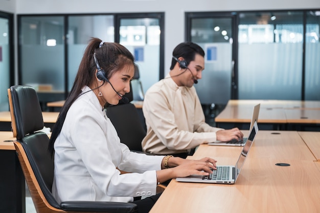 Przyjazny operator azjatycka kobieta agent z zestawem słuchawkowym pracującym z laptopem na konsultacje z klientem w biurze z kolegą w miejscu pracy