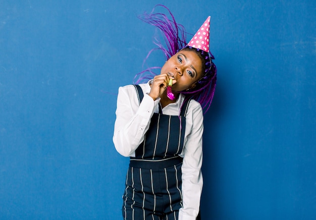 Przyjęcie Urodzinowe, Karnawał Nowy Rok. Młoda Uśmiechnięta Afrykańska Kobieta świętuje Jasne Wydarzenie Na Niebieskiej Przestrzeni, Nosi Elegancką Modną Białą Spódnicę I Czarne Spodnie, Z Różowym Imprezowym Kapeluszem Z Noisemaker.